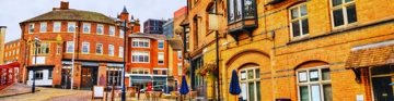 Orange-brick buildings and a street scene with shopfronts, signs, and closed blue parasols.