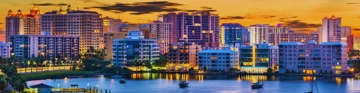 Waterfront city skyline with tall buildings reflected in calm water at sunset.