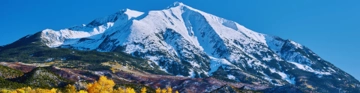 Snow-covered mountain rising above colourful woodland beneath a clear blue sky.